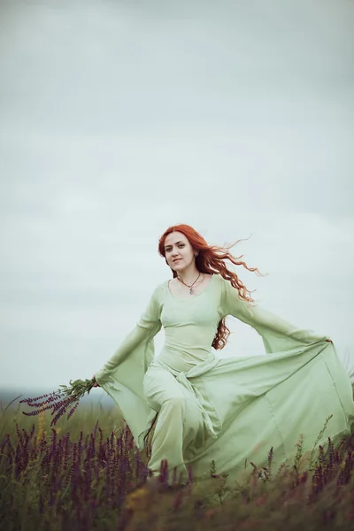 Chica pelirroja joven en vestido medieval caminando por el campo con flores de salvia. Concepto de viento. Fantasía —  Fotos de Stock