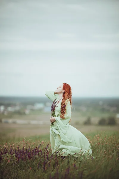 Chica pelirroja joven en vestido medieval caminando por el campo con flores de salvia. Concepto de viento. Fantasía — Foto de Stock