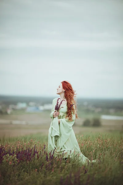 Chica pelirroja joven en vestido medieval caminando por el campo con flores de salvia. Concepto de viento. Fantasía —  Fotos de Stock
