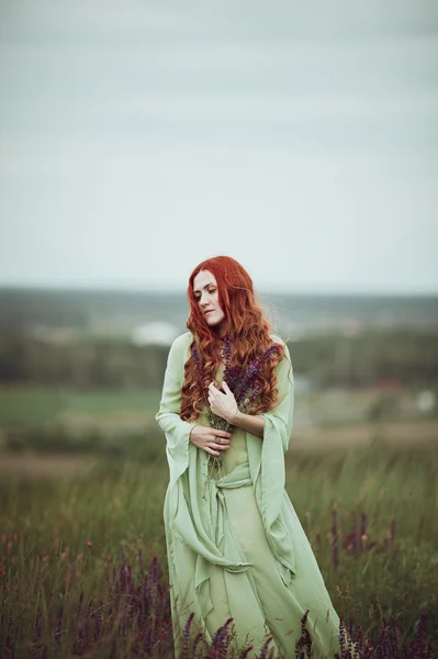 Junges rothaariges Mädchen in mittelalterlichem Gewand, das mit Salbeiblüten durch das Feld geht. Windkonzept. Fantasie — Stockfoto