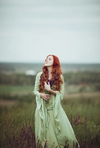 Jeune fille rousse en robe médiévale marchant à travers le champ avec des fleurs de sauge. Concept de vent. Fantaisie — Photo