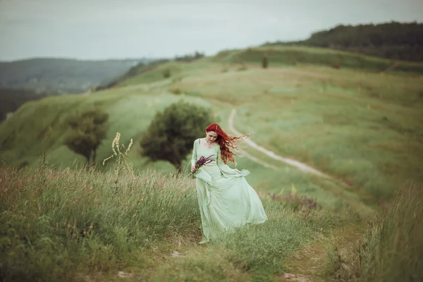 Chica pelirroja joven en vestido medieval caminando por el campo con flores de salvia. Concepto de viento. Fantasía — Foto de Stock