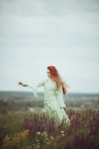 Junges rothaariges Mädchen in mittelalterlichem Gewand, das mit Salbeiblüten durch das Feld geht. Windkonzept. Fantasie — Stockfoto