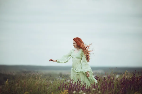 Chica pelirroja joven en vestido medieval caminando por el campo con flores de salvia. Concepto de viento. Fantasía — Foto de Stock