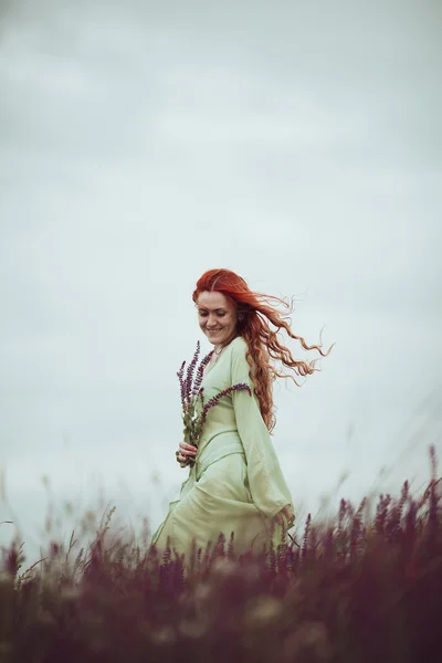 Jeune fille rousse en robe médiévale marchant à travers le champ avec des fleurs de sauge. Concept de vent. Fantaisie — Photo