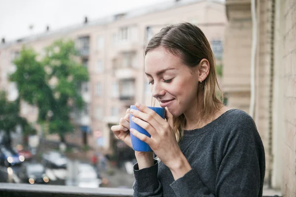 Beautiful young girl with a cup of morning coffee standing on the balcony in the rain. Thinking concept. — Stock Photo, Image