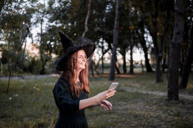 Koyu renk elbiseli ve cadı şapkalı güzel bir kadın sosyal ağlar için selfie çekiyor. Cadılar Bayramı partisi kostümü. Orman, sonbahar ağaçlarının olduğu park.