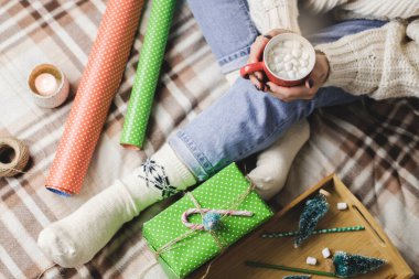 Genç bir kadın, rahat, yün, beyaz bir kazakla ekose üzerinde oturur ve elinde lokum dolu bir fincan kakao tutar. Hygge Yeni Yıl, tatil hazırlıkları. Mumlar, Noel baloları, hediyeler, ambalaj kağıtları..