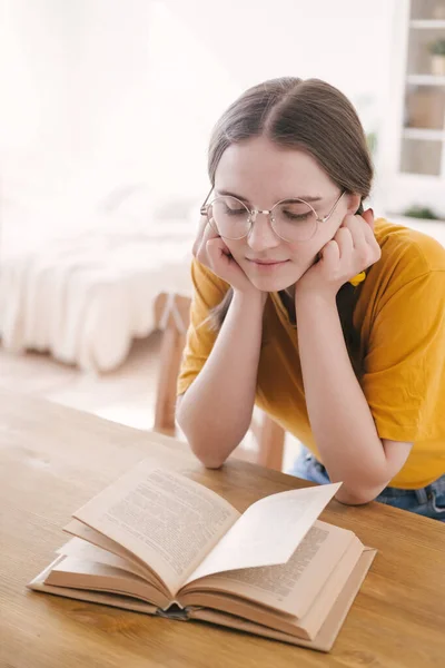 Ung vacker kvinna student i orange T-shirt och glasögon läser bok sitter vid bordet. Förberedelser inför tentamen, hemundervisning i självisolering. — Stockfoto