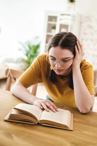 Ung vacker kvinna student i orange T-shirt och glasögon läser bok sitter vid bordet. Förberedelser inför tentamen, hemundervisning i självisolering. — Stockfoto