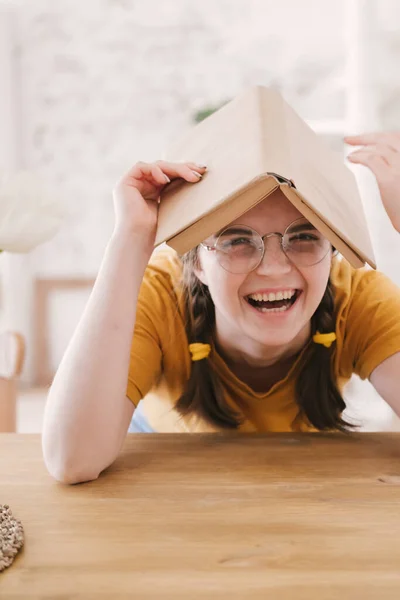 Jonge mooie studente in oranje T-shirt en bril leest boek zittend aan tafel. Voorbereiding van examens, thuis studeren over zelfisolatie. — Stockfoto