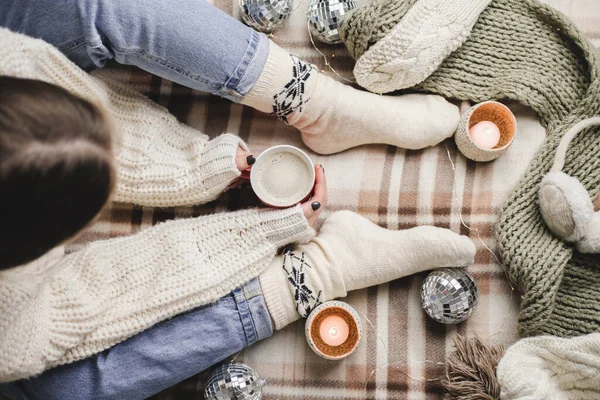 Young woman sits on plaid in cozy knitted woolen white sweater and socks holds cup of cocoa in her hands. Hygge New Year, cozy Christmas, preparation for holidays. Candles, Christmas balls. — Stock Photo, Image