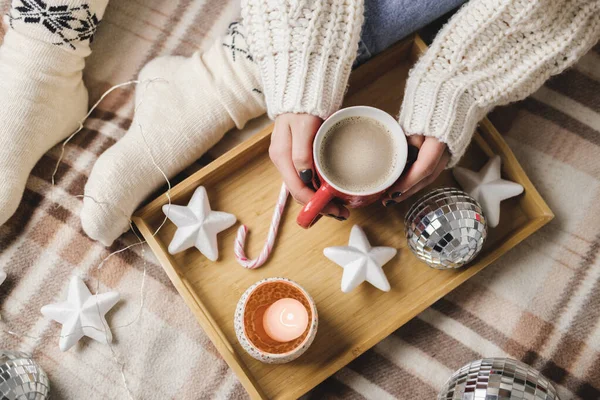 Young woman sits on plaid in hugge cozy knitted woolen white sweater and holds cup of cocoa in her hands. Wooden tray with mug of chocolate with toy tree, candle, stars. — Stock Photo, Image