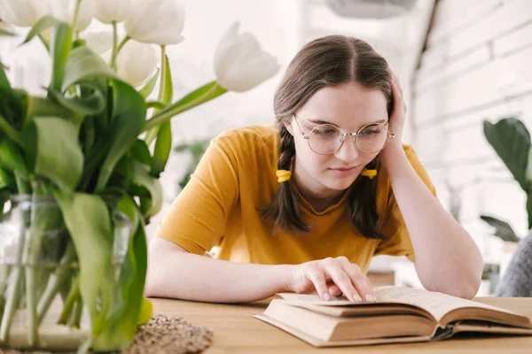 Ung vacker kvinna student i orange T-shirt och glasögon läser bok sitter vid bordet. Förberedelser inför tentamen, hemundervisning i självisolering. — Stockfoto