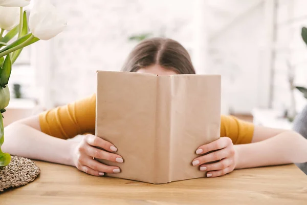 Ung vacker kvinna student i orange T-shirt och glasögon läser bok sitter vid bordet. Förberedelser inför tentamen, hemundervisning i självisolering. — Stockfoto