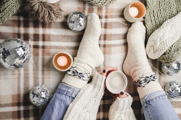 Mujer joven se sienta a cuadros en suéter blanco de lana de punto acogedor y calcetines sostiene la taza de cacao en sus manos. Hygge Año Nuevo, Navidad acogedora, preparación para las vacaciones. Velas, bolas de Navidad. — Foto de Stock