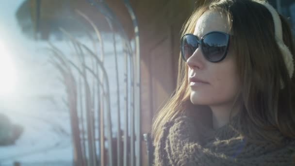 Mujer joven en auriculares de piel, gafas de sol y abrigo de color turquesa, bufanda y con mochila disfruta del sol y el clima. Alrededor de la estación de esquí de invierno, nieve, montañas, ascensores. Movimiento lento. — Vídeos de Stock