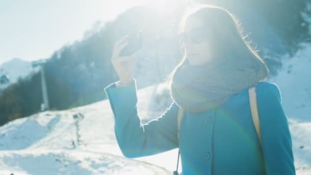 Jonge vrouw in bont koptelefoon, zonnebril en turquoise jas, sjaal en met rugzak neemt foto of video op smartphone. Rond het skigebied, sneeuw, bergen, liften. Langzame beweging. — Stockvideo
