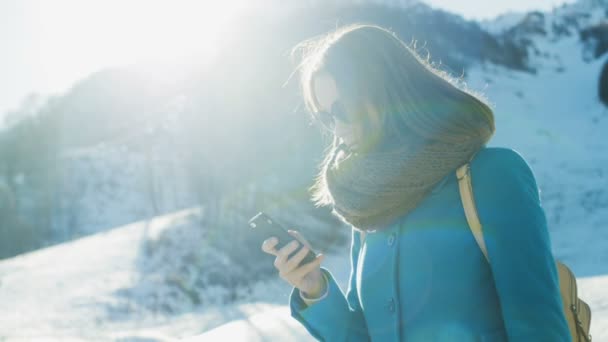 Giovane donna in pelliccia cuffie, occhiali da sole e cappotto turchese, sciarpa e con zaino utilizza il social network nello smartphone. Intorno alla stazione sciistica invernale, neve, montagne, impianti di risalita. Rallentatore. — Video Stock