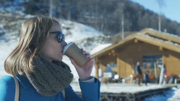 Mujer joven en auriculares de piel, gafas de sol y abrigo turquesa, bufanda y con mochila, bebe café de taza de papel. Alrededor de la estación de esquí de invierno, nieve, montañas, ascensores. Movimiento lento. — Vídeos de Stock