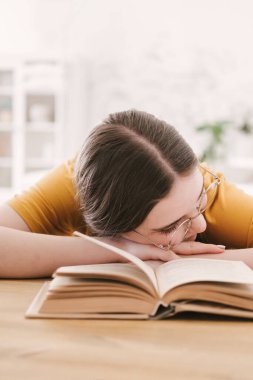 Turuncu tişörtlü ve gözlüklü genç bayan öğrenci masada oturmuş kitap okuyor. Sınavlara hazırlanmak, evde tek başına çalışmak..