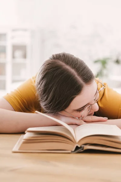 Ung vacker kvinna student i orange T-shirt och glasögon läser bok sitter vid bordet. Förberedelser inför tentamen, hemundervisning i självisolering. — Stockfoto