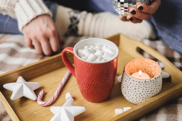 La jeune femme s'assoit sur le plaid dans un confortable pull en laine blanche et tient une tasse de cacao avec des guimauves. Hygge bannière Nouvel An, une préparation confortable pour les vacances. Bougies, boules de Noël dans un plateau en bois. — Photo