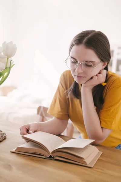 Ung vacker kvinna student i orange T-shirt och glasögon läser bok sitter vid bordet. Förberedelser inför tentamen, hemundervisning i självisolering. — Stockfoto