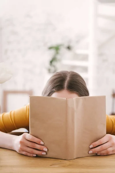 Turuncu tişörtlü ve gözlüklü genç bayan öğrenci masada oturmuş kitap okuyor. Sınavlara hazırlanmak, evde tek başına çalışmak.. — Stok fotoğraf