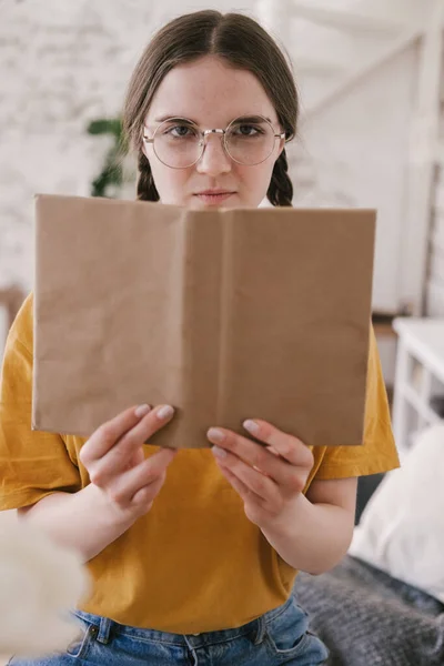 Ung vacker kvinna student i orange T-shirt och glasögon läser bok sitter vid bordet. Förberedelser inför tentamen, hemundervisning i självisolering. — Stockfoto