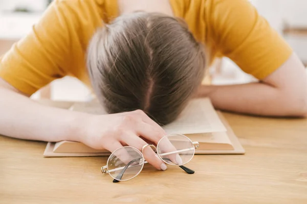 Молода красива студентка в помаранчевій футболці і окулярах читає книгу, сидячи за столом. Підготовка до іспитів, навчання вдома на самоізоляції . — стокове фото