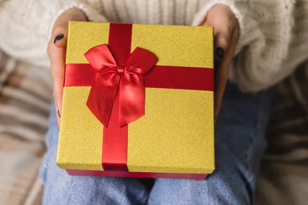 Mujer joven se sienta en la cama en un acogedor suéter blanco de lana de punto y la celebración de la caja de regalo con oro rojo. Hygge, Año Nuevo, Navidad, preparativos navideños. Velas, bolas de Navidad, regalos, malvaviscos de cacao. — Foto de Stock