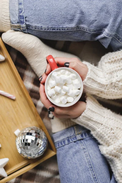 Молодая женщина сидит на клетке в уютном вязаном шерстяном белом свитере и держит чашку какао с зефиром. Новый год, уютная подготовка к праздникам. Свечи, рождественские шары в деревянном подносе. — стоковое фото
