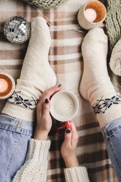 Młoda kobieta siedzi na kratę w przytulnej dzianiny wełniany biały sweter i skarpety trzyma filiżankę kakao w rękach. Hygge Nowy Rok, przytulne Boże Narodzenie, przygotowanie do wakacji. Świece, świąteczne kulki. — Zdjęcie stockowe