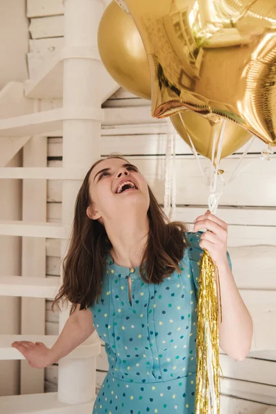 Молода красива дівчина в блакитній сукні з білими поляками відзначає свій день народження і насолоджується золотими повітряними кулями. День народження вдома під час самоізоляції . — стокове фото
