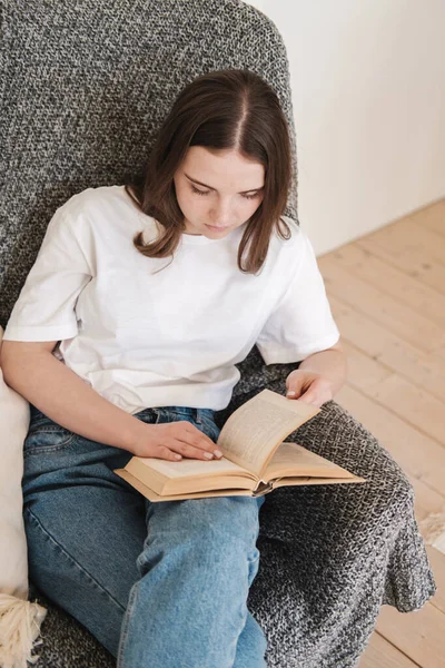 Ung kvinna i jeans och vit T-shirt sitter i grå fåtölj och läser pappersbok. Begreppet avkopplande semester, hem komfort, hygge, långsamt boende — Stockfoto