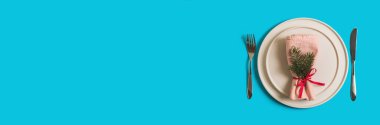 Noel ve Yeni Yıl kutlamaları için içi boş tabak ve çatal bıçak dolu servis masası. Tabakta Noel ağacı dallı peçete var. Parlak mavi arkaplanda yassı pankart. Üst görünüm