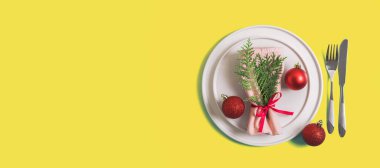 Noel ve Yeni Yıl kutlamaları için servis tabağı ve çatal bıçak takımı olan web sitesi afişi. Tabakta Noel ağacı dallı peçete ve kırmızı toplar var. Aydınlatıcı sarı arkaplan üzerine düz çizgi. Üst görünüm.
