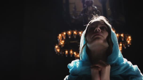Young millennial woman in pink dress and blue headscarf stands in church, temple and prays, looks at the camera with a serious expression. Slow motion. — Stock Video
