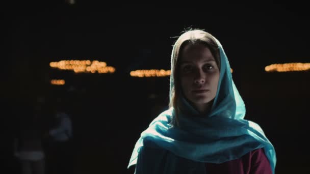 Die junge Millennials-Frau in rosa Kleid und blauem Kopftuch steht in Kirche, Tempel und betet, blickt mit ernstem Gesichtsausdruck in die Kamera. Zeitlupe. — Stockvideo