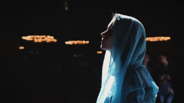 Young millennial woman in pink dress and blue headscarf stands in church, temple and prays, looks at the camera with a serious expression. Slow motion. — Stock Video