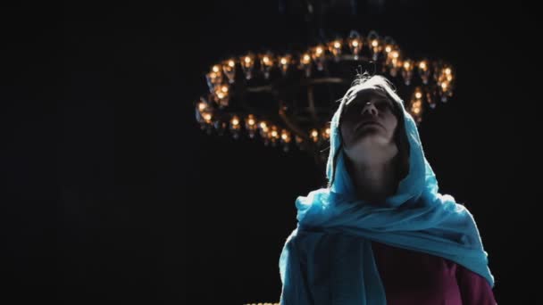 Young millennial woman in pink dress and blue headscarf stands in church, temple and prays, looks at the camera with a serious expression. Slow motion. — Stock Video