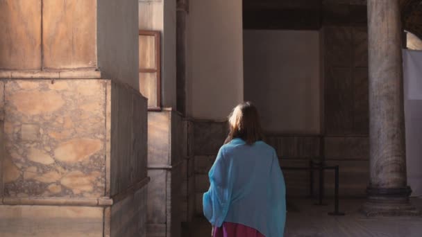 ピンクのドレスと青いスカーフの若い千年紀の女性は、真剣な表情でカメラを見て、部屋を前後に窓の外を歩く。スローモーション. — ストック動画