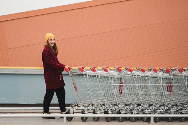 身穿勃艮第外套头戴黄色帽子的年轻漂亮的女人走在超市停车场的屋顶上，坐着手推车. — 图库照片