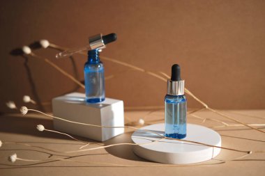 Bir şişe serum, beyaz yuvarlak ve dikdörtgen bir kürsüde kurumuş çiçek dallarıyla duruyor. Wabisabi tarzı, toprak rengi. Cilt Kozmetik Düzenleme.
