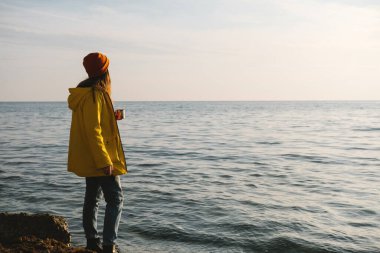 Parlak sarı paltolu genç bir kadın deniz kıyısında ya da okyanusta çay, kahve ve manzaranın tadını çıkarıyor. Özgürlük kavramı, düşüncelilik, farkındalık..