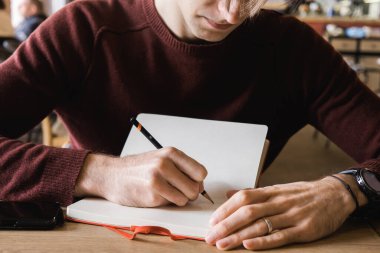 Genç bir adam kafede oturur, kahve içer ve not defterine notlar ve çizimler yazar..