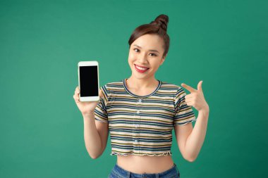 Gülümseyen Asyalı genç bir kadın işaret parmağıyla yeşil arka planda izole edilmiş boş ekran telefonunu işaret ediyor..
