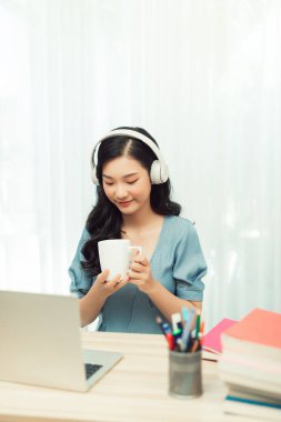 Gözlüklü, kablosuz kulaklıklı, kameraya gülümseyen ve elinde kupayla dizüstü bilgisayarla oturan bağımsız bir kadın. 