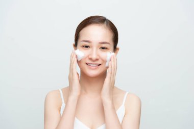 Tatmin olmuş güzel bir kadın yüz masajı yapıyor, beyaz köpüğe sahip, cilt bakımlı, gülümseyen, beyaz stüdyo duvarının üstünde mankenler.